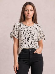 Model wearing an ivory printed boat neck blouse with black abstract pattern, short tied sleeves, and a relaxed straight silhouette, paired with black jeans.