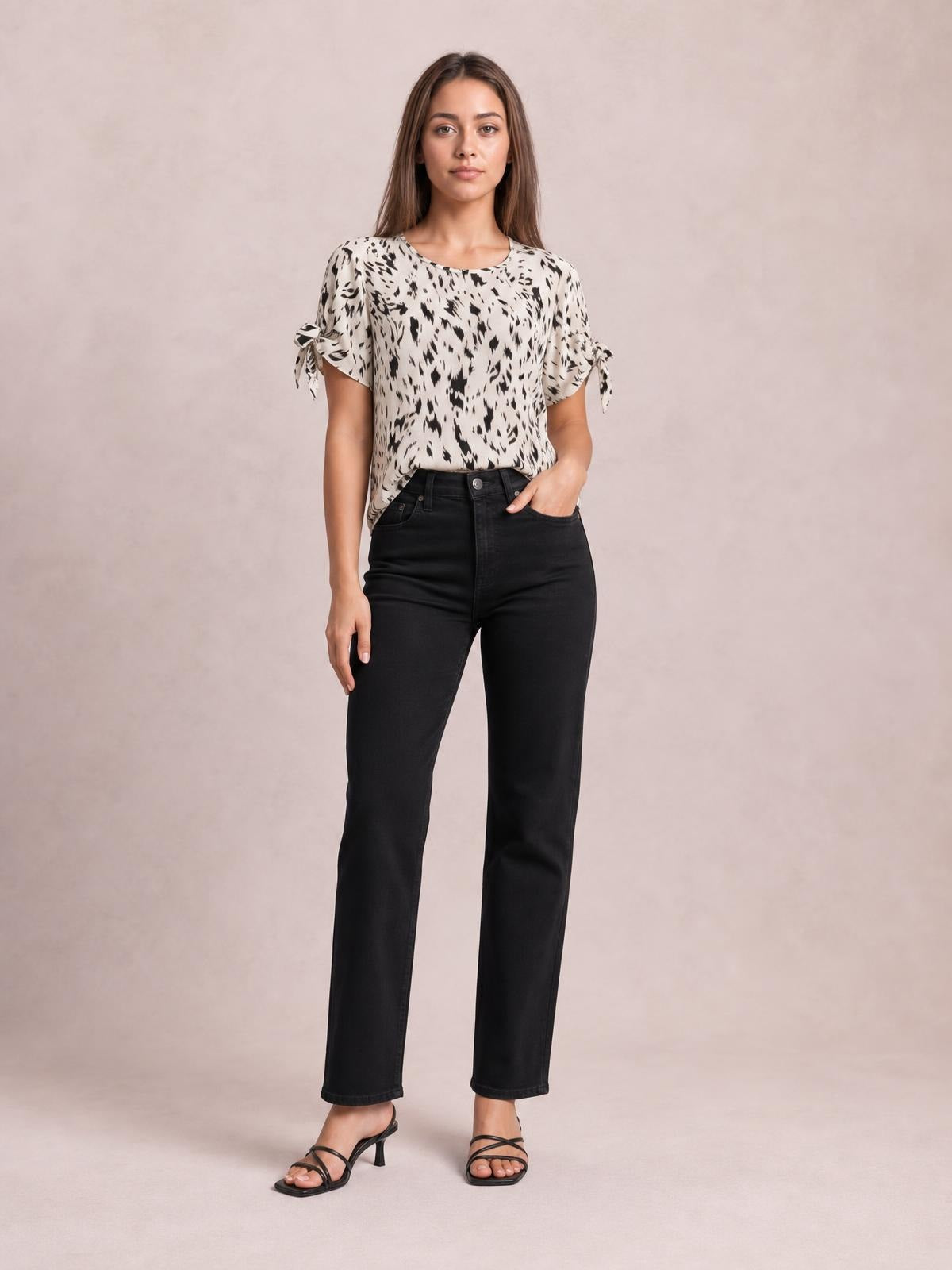 Model stands in a cream and black printed boat neck blouse with short sleeves, tied bow details, and a relaxed silhouette, paired with black straight-leg jeans.