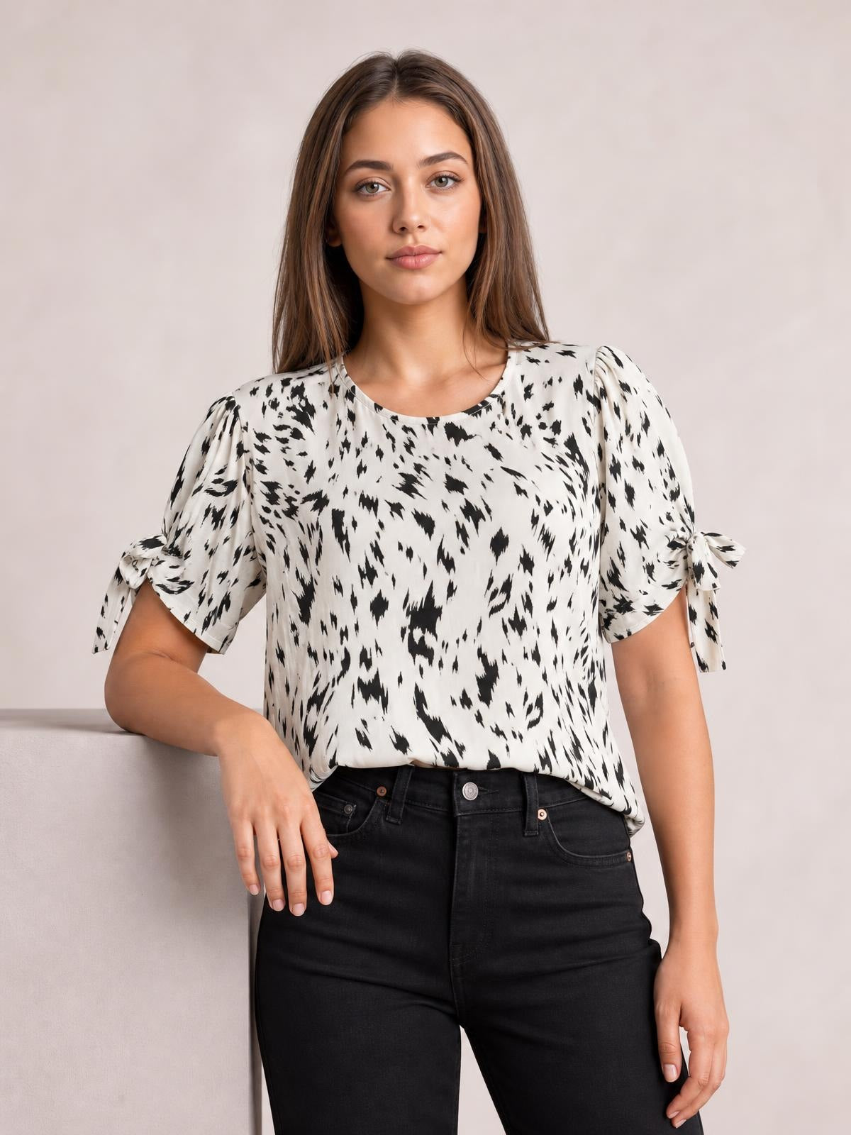 Model wearing a black-and-white printed boat neck blouse with a relaxed silhouette, puff sleeves, and tied sleeve details, posed with one hand resting on a ledge.
