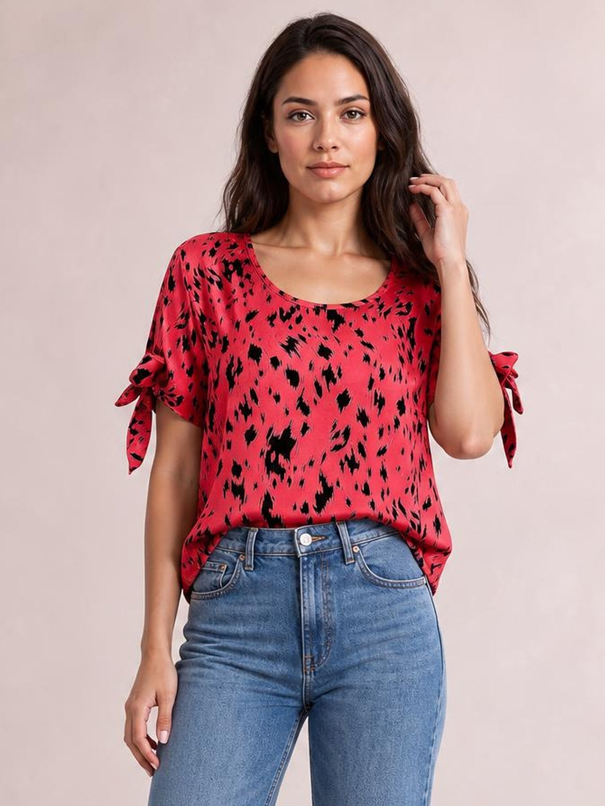Model wearing a loose red printed boat neck blouse with tied short sleeves, paired with blue jeans, posing with one hand touching her hair.
