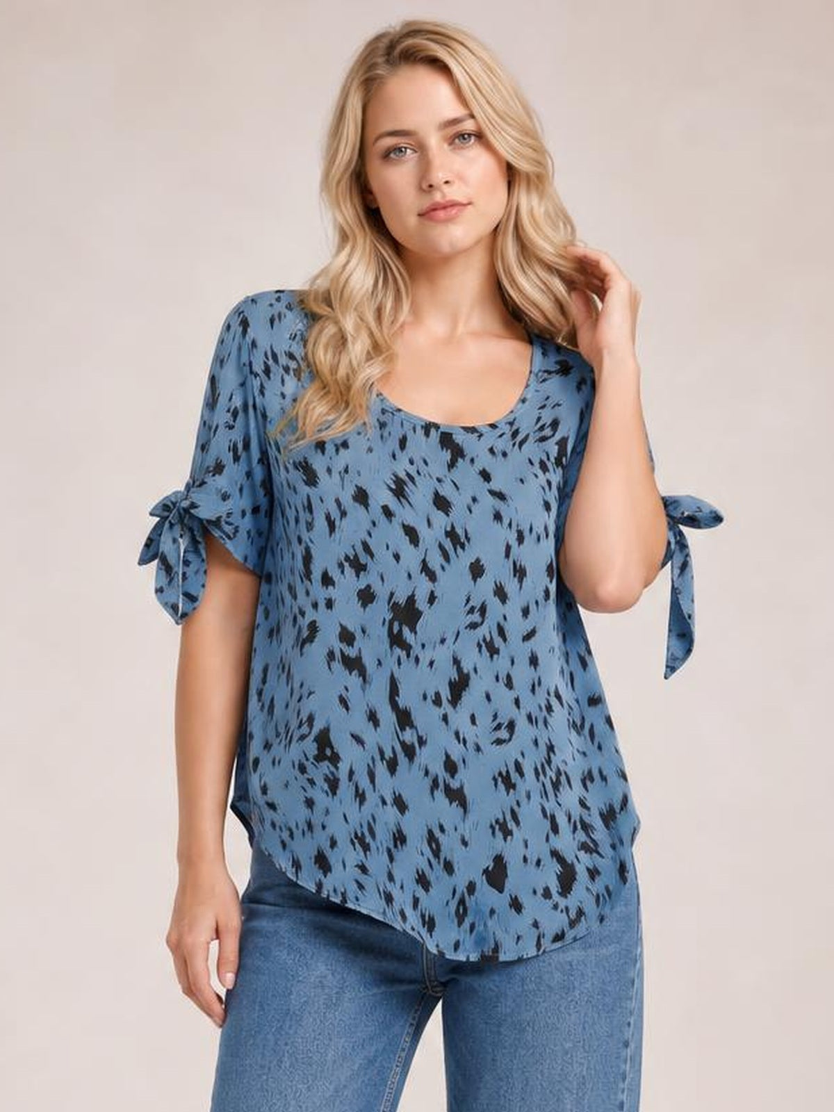 A model poses in a blue printed blouse with a wide boat neckline, loose silhouette, short sleeves, and tied bow details at the cuffs.