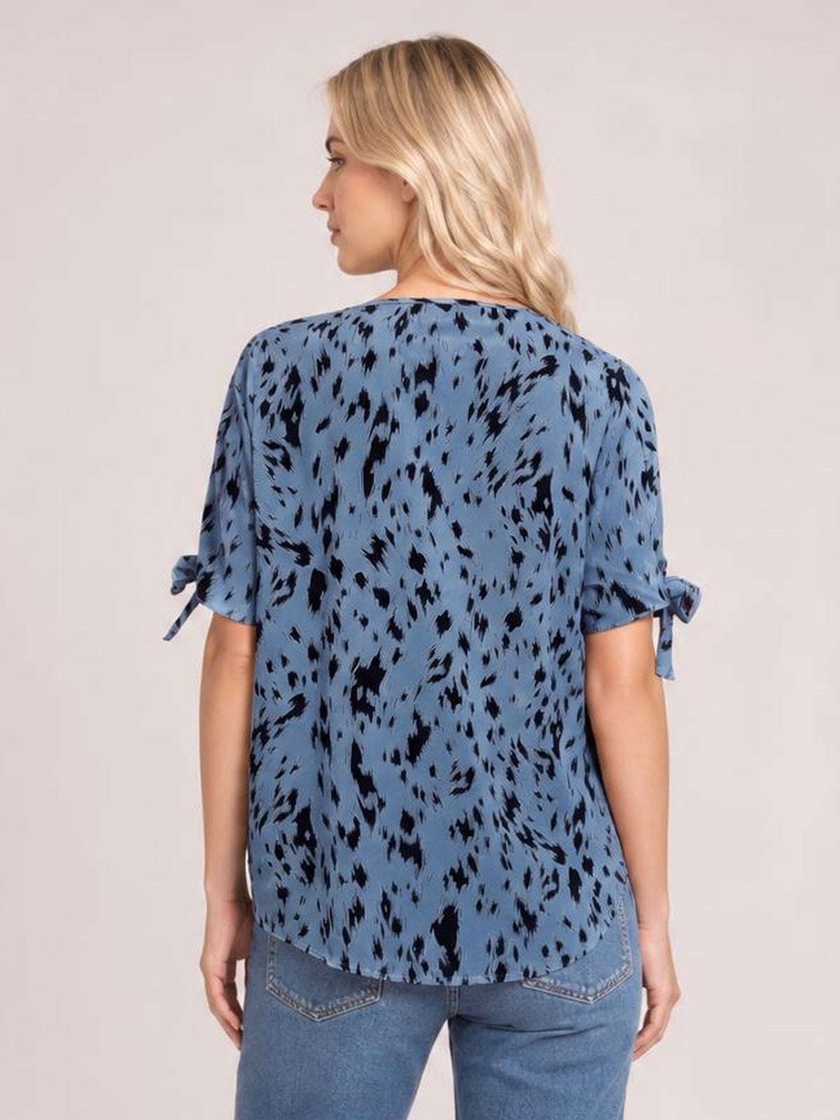 Model shown from behind wears a loose blue printed boat neck blouse with black patterning, short sleeves, tied details, and a curved hem.