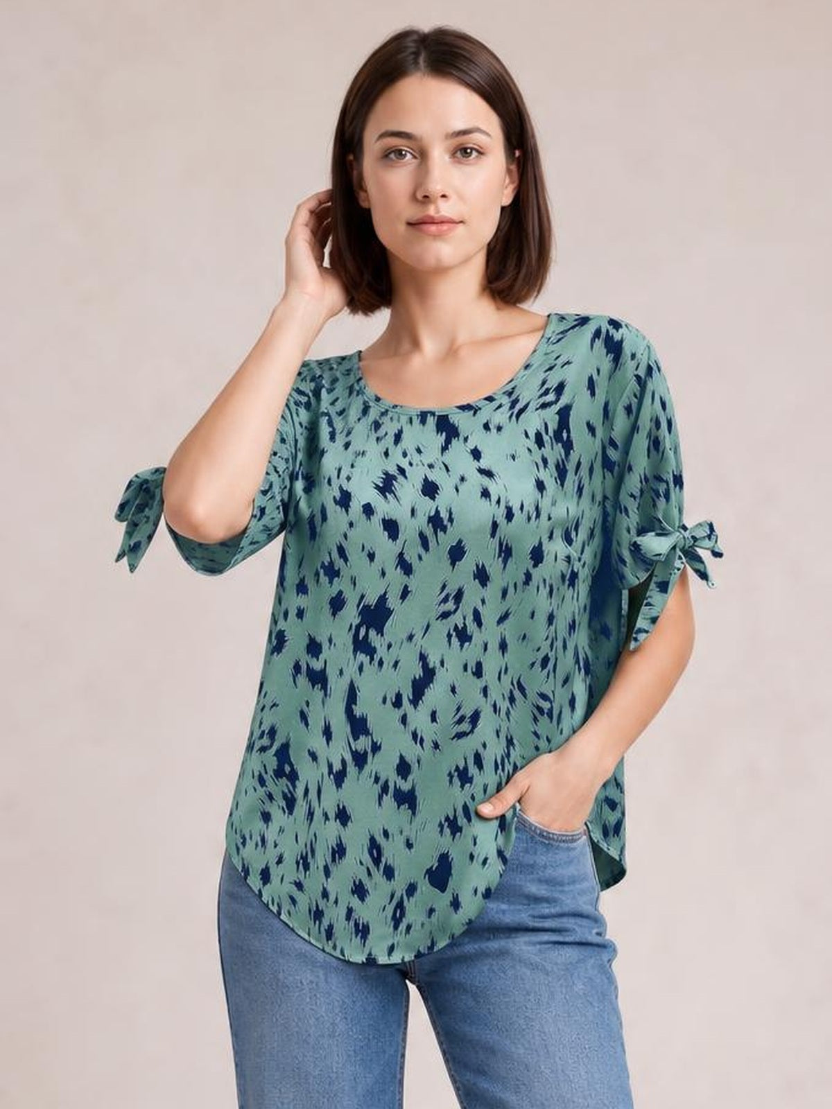 A model wears a loose sage green printed boat neck blouse with navy accents, tie-detail three-quarter sleeves, and a curved hem while posing with one hand in her jeans pocket.