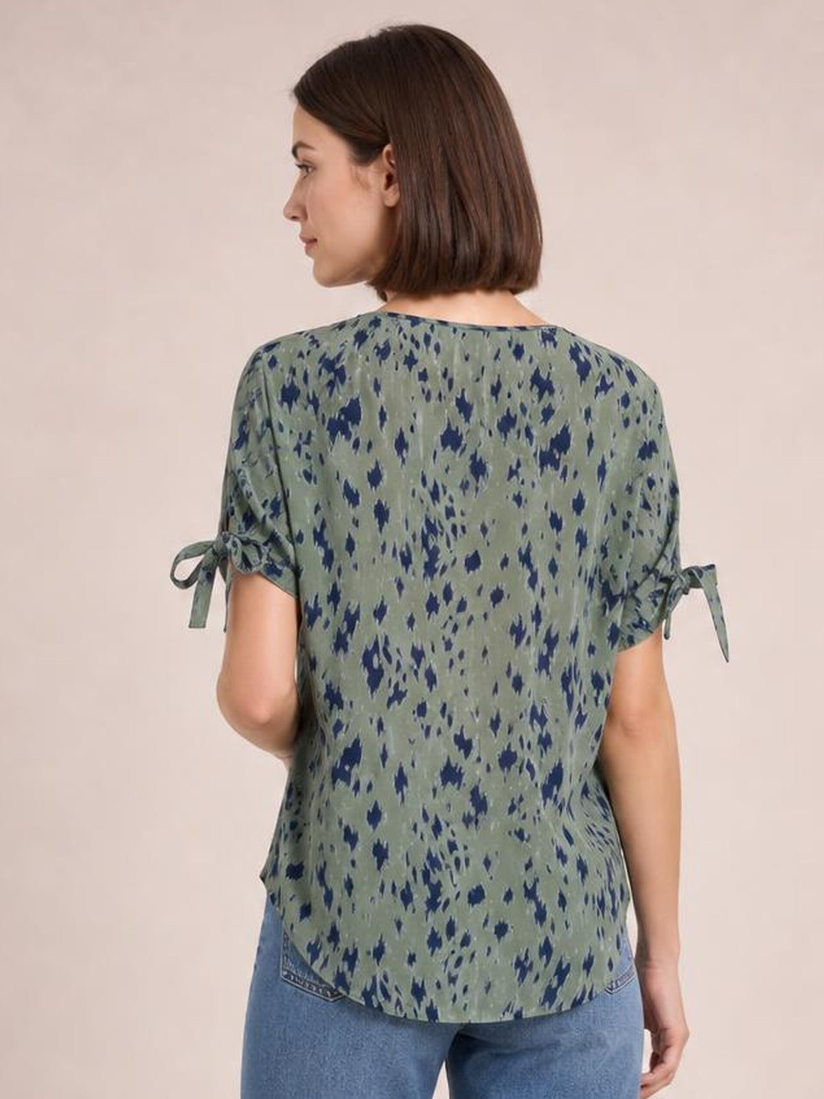 A model wears a relaxed olive-green printed boat neck blouse with navy abstract spots, tied short sleeves, and a curved hem while turned to the side.