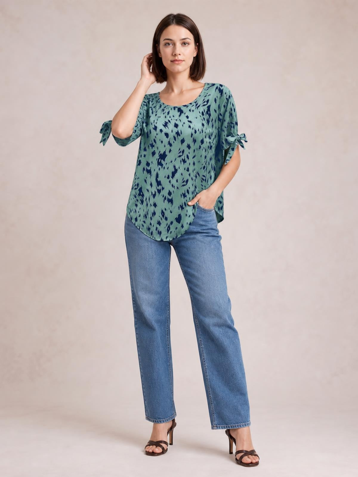 A model stands in a relaxed pose wearing a mint-green and navy printed boat-neck blouse with tied short sleeves and a loose, curved-hem silhouette.