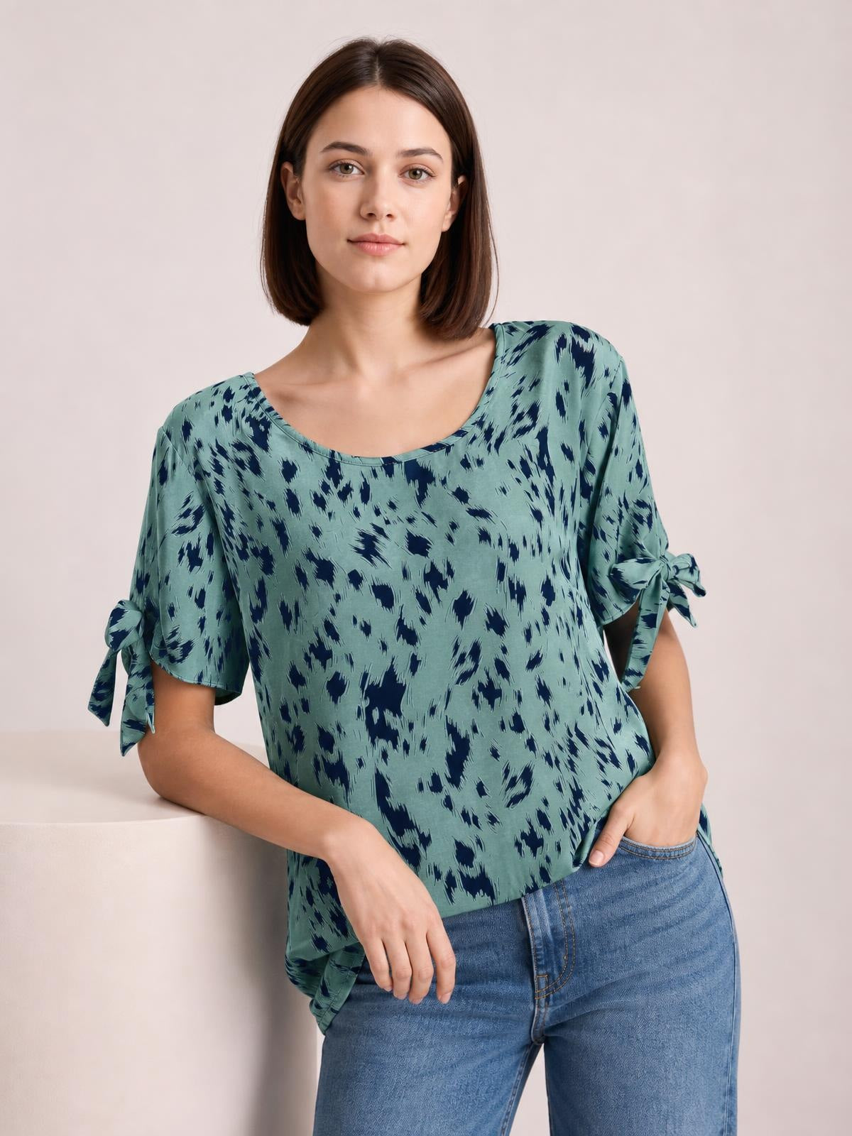 Model poses in a teal and navy printed boat neck blouse with a relaxed silhouette, short sleeves tied at the cuffs, and a loose untucked hem.