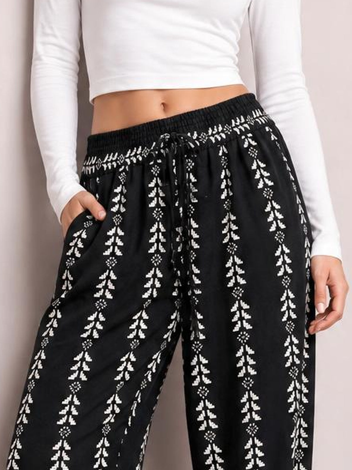 A cropped model wears black printed high waist pants with white vertical motifs, an elastic waistband, front tie, relaxed wide-leg silhouette, and side pockets.
