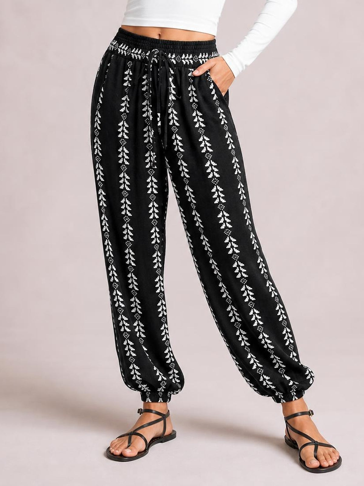 A model poses in black printed high-waist pants with a tied drawstring, relaxed wide-leg silhouette, vertical white motifs, side pockets, and elastic cuffs.