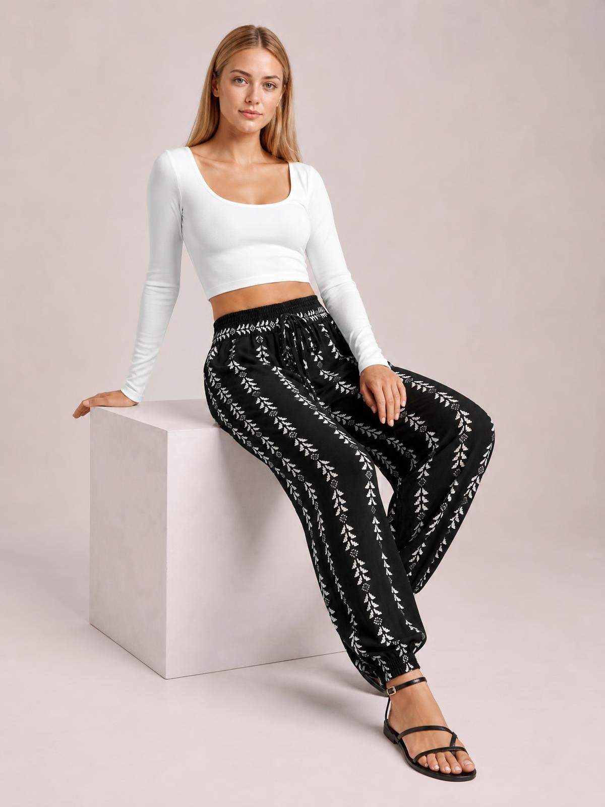 A seated model wears black printed high-waist pants with a tied drawstring waistband and relaxed wide legs, posed with one leg extended.