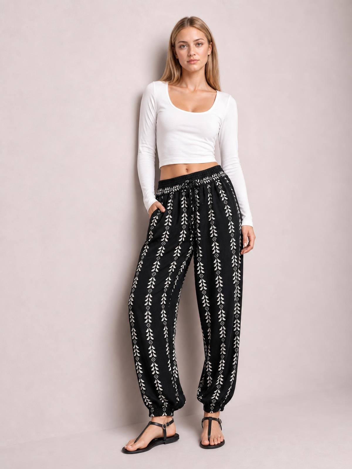 Model wearing black printed high waist pants with a tied drawstring waist, relaxed tapered legs, and elasticized ankle cuffs, posed with one hand in a pocket.