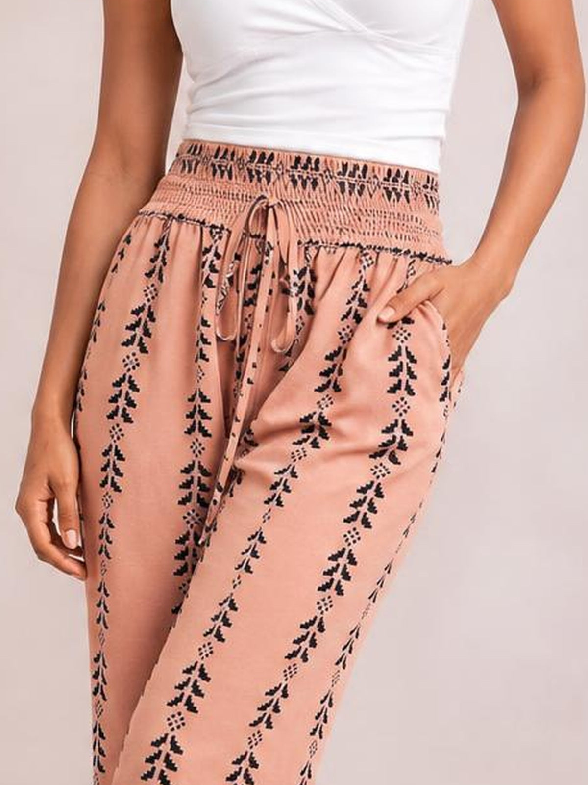 A model wears peach-pink printed high-waist pants with a smocked waistband, front tie, relaxed silhouette, and black geometric floral motifs.