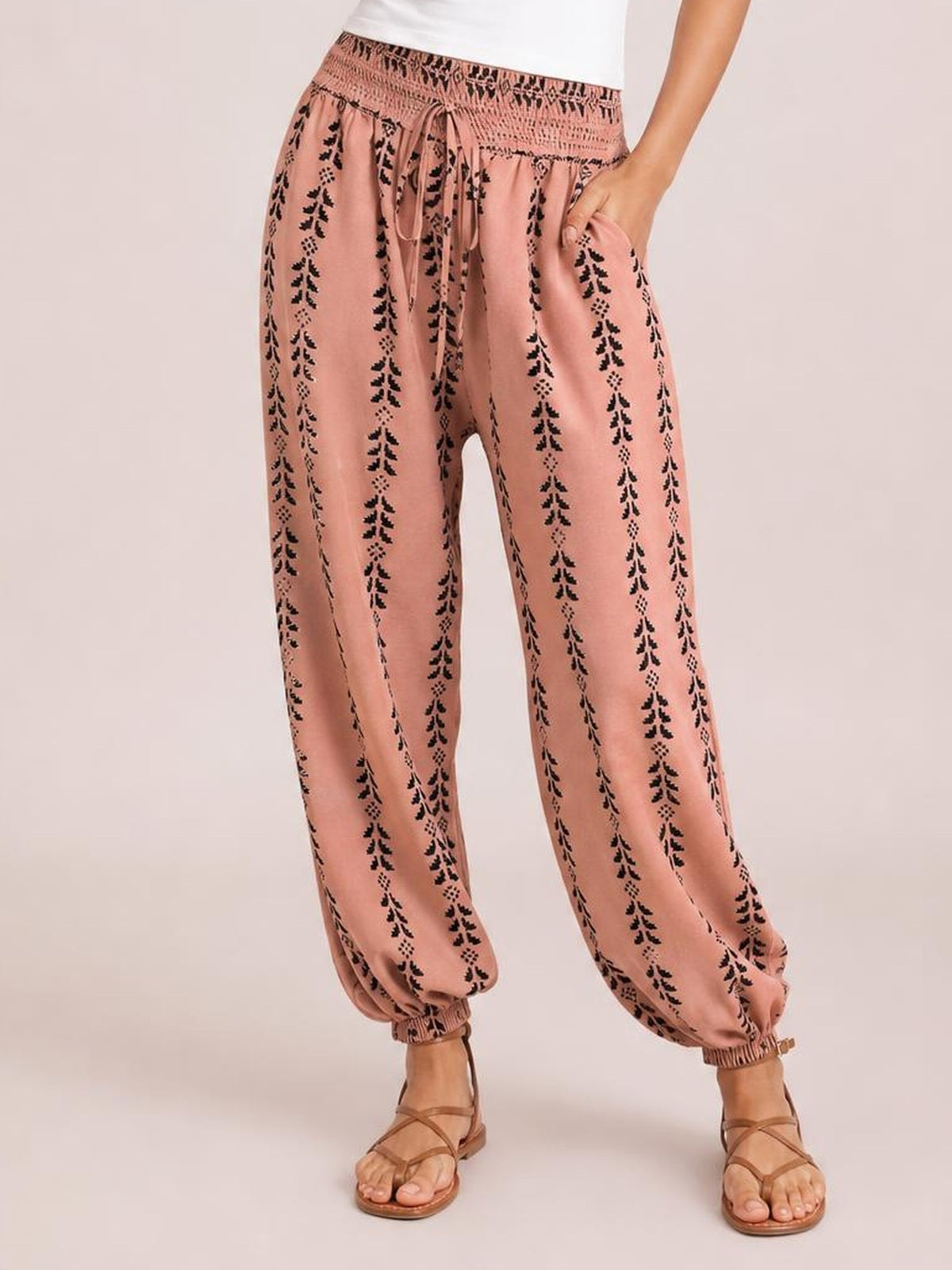 A model stands with one hand in a pocket wearing dusty pink high-waist pants with black vertical print, a tie waist, wide legs, and elastic cuffs.