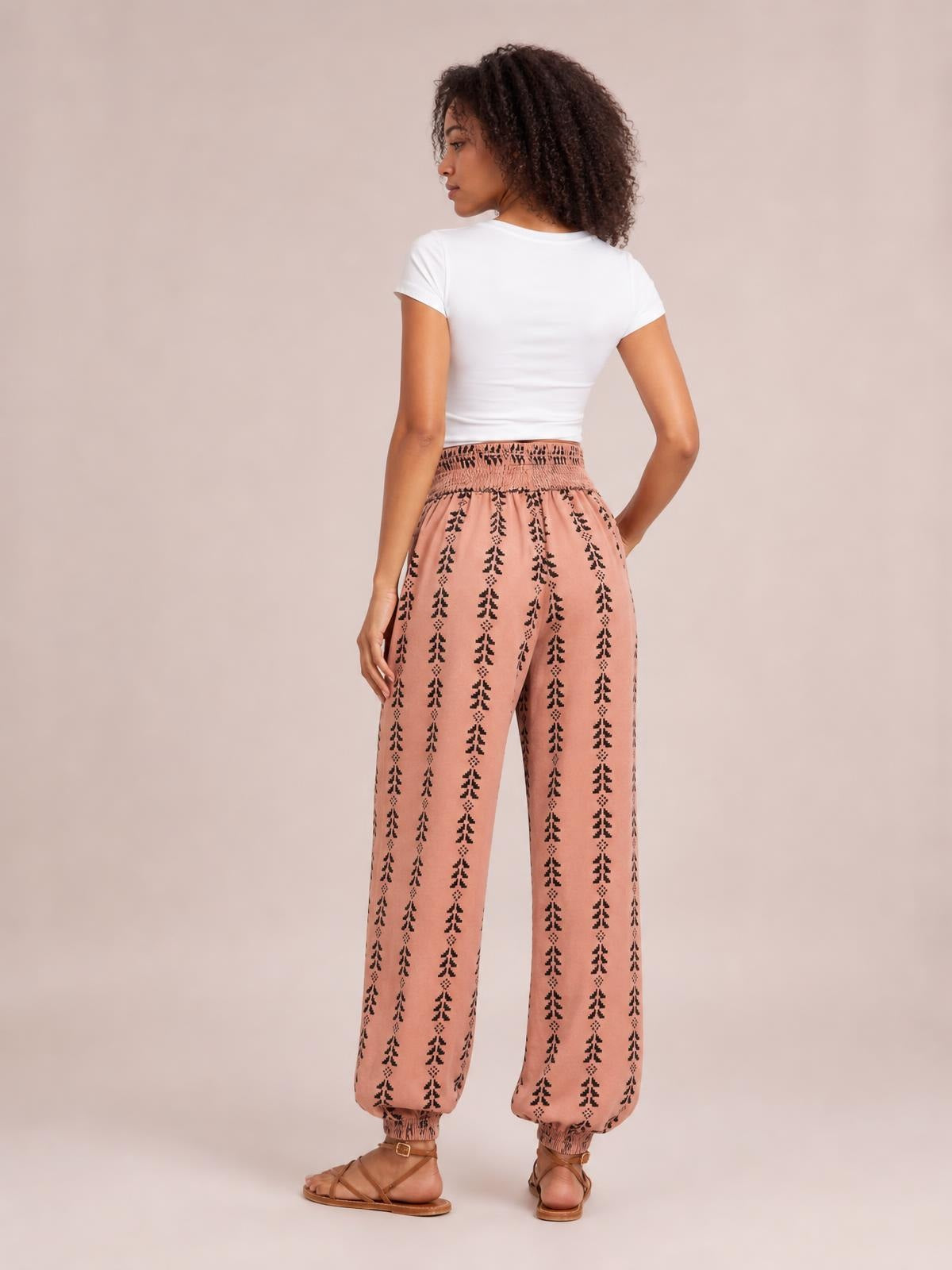 A model poses from the back in pink printed high-waist pants with a smocked waistband, relaxed wide-leg silhouette, and gathered ankle cuffs.