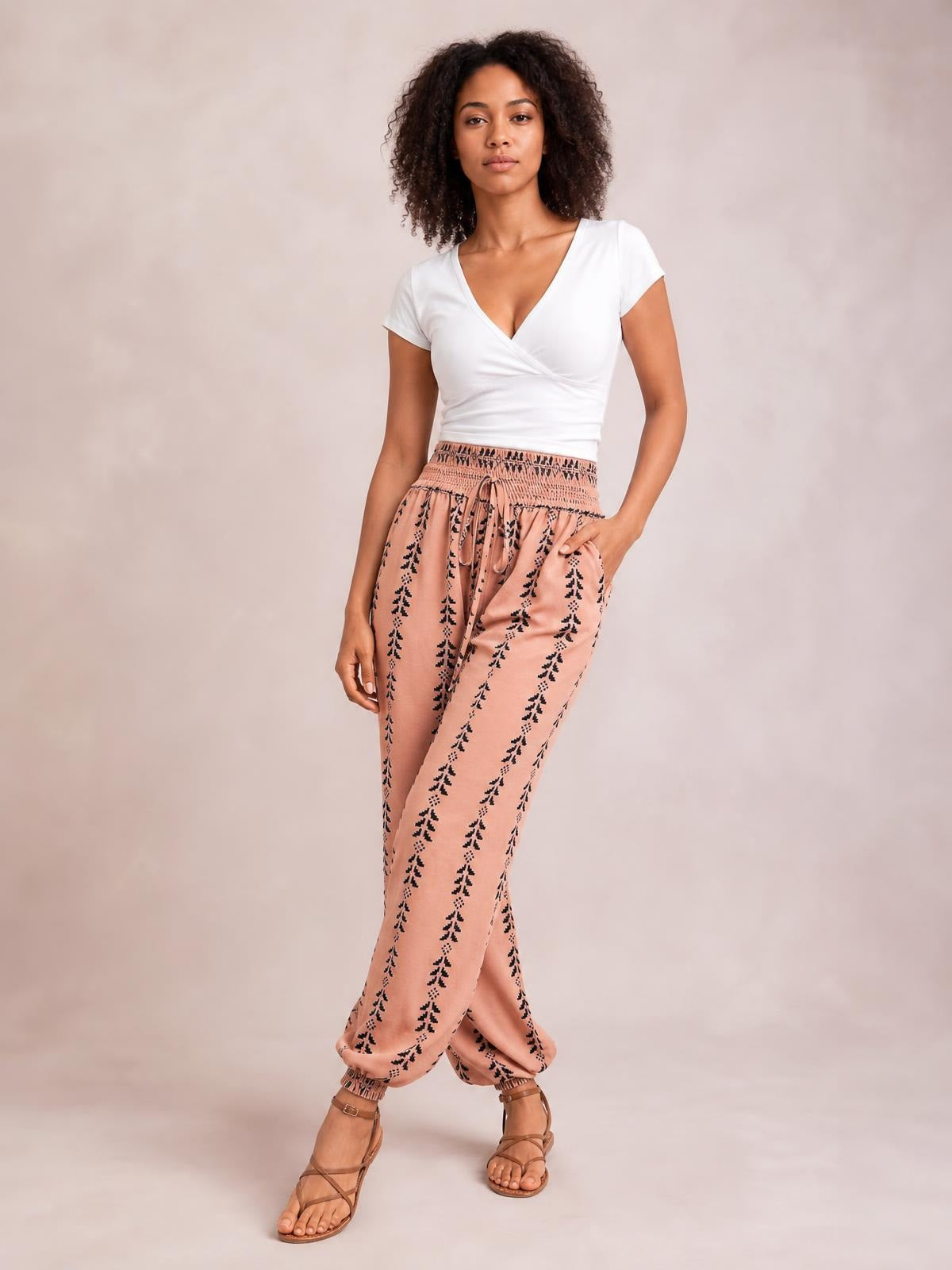 A model poses in pink high-waist printed pants with black motifs, a tied drawstring waist, relaxed tapered legs, and gathered ankle cuffs.