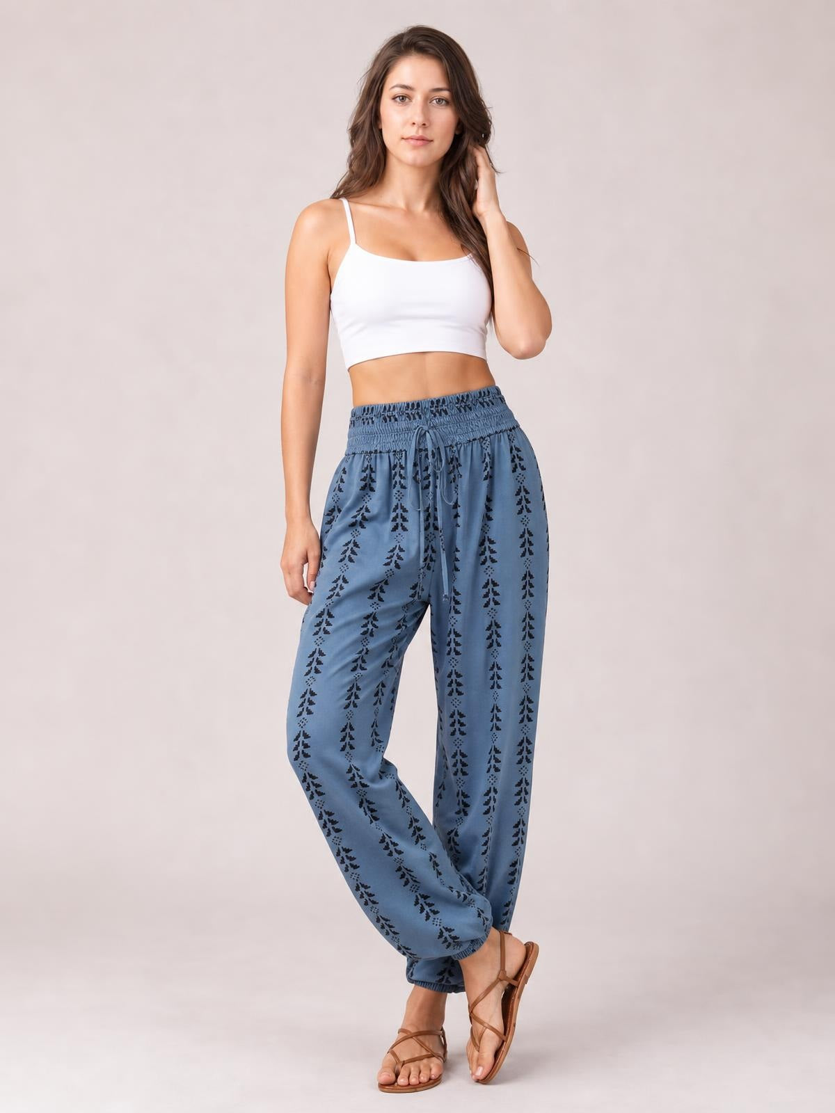 Model poses in blue printed high-waist pants with a tied drawstring waist, smocked waistband, relaxed tapered silhouette, and elastic cuffs.
