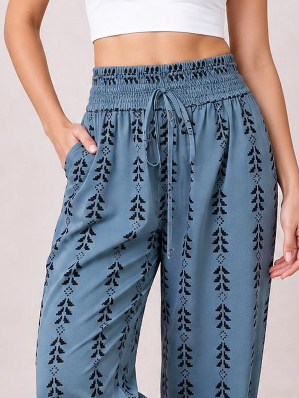 A cropped model wears blue tied printed high waist pants with a smocked waistband, front drawstring, black geometric pattern, and relaxed wide-leg silhouette.