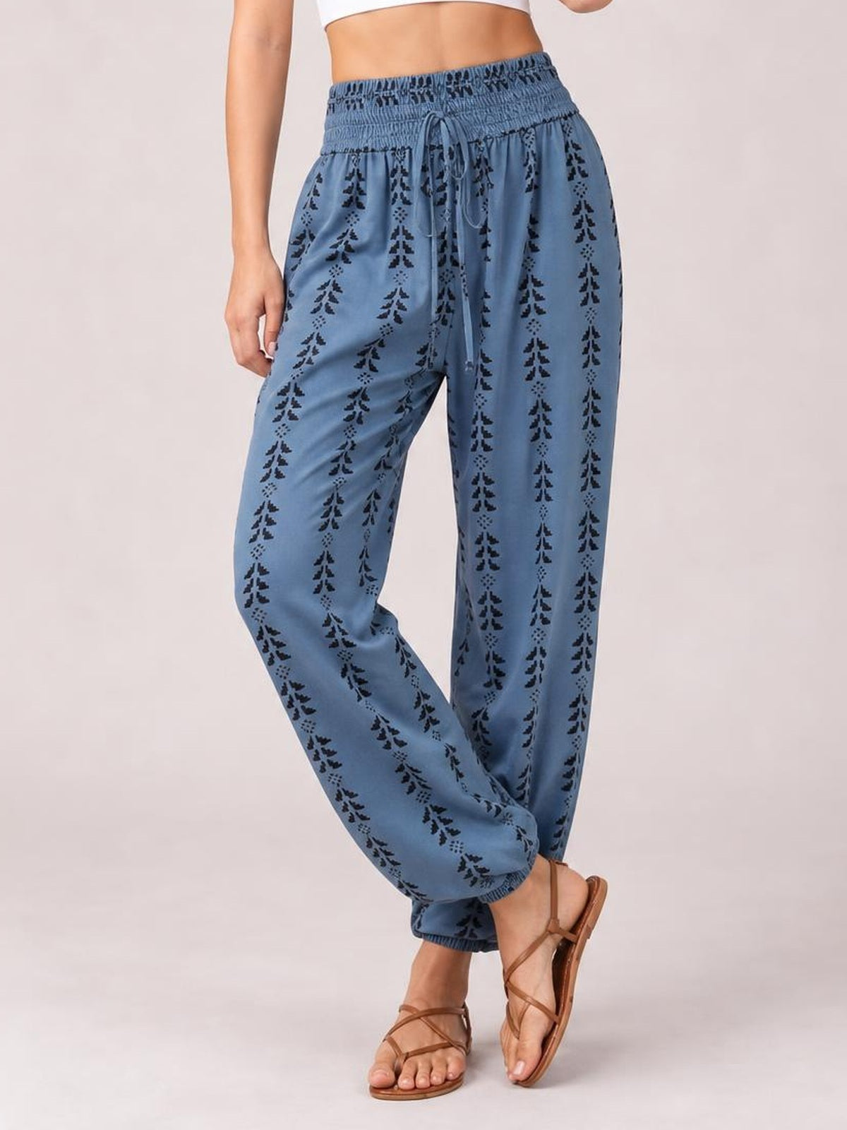 Model wearing blue printed high-waist pants with a tied drawstring waist, smocked waistband, relaxed tapered legs, and elastic cuffs while crossing her ankles.
