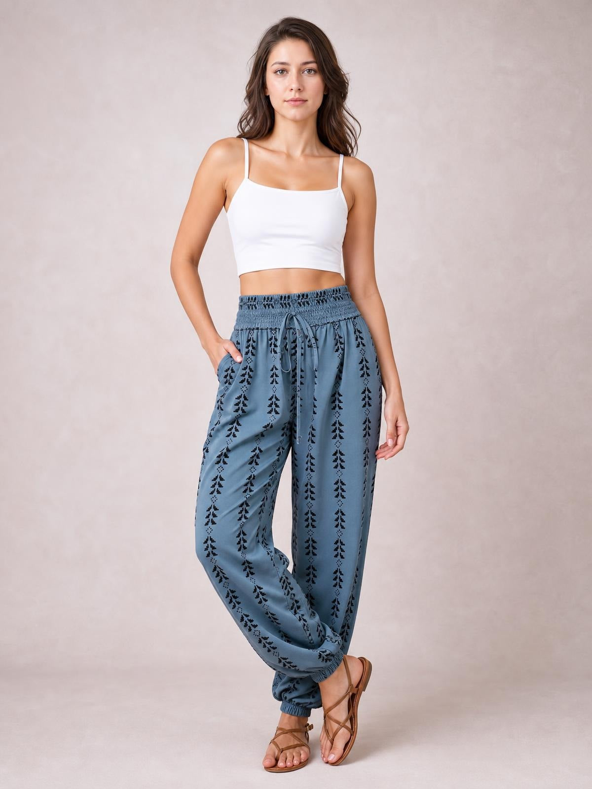 A model poses in blue printed high-waist pants with black motifs, a smocked drawstring waistband, relaxed wide legs, elastic cuffs, and a hand in the pocket.