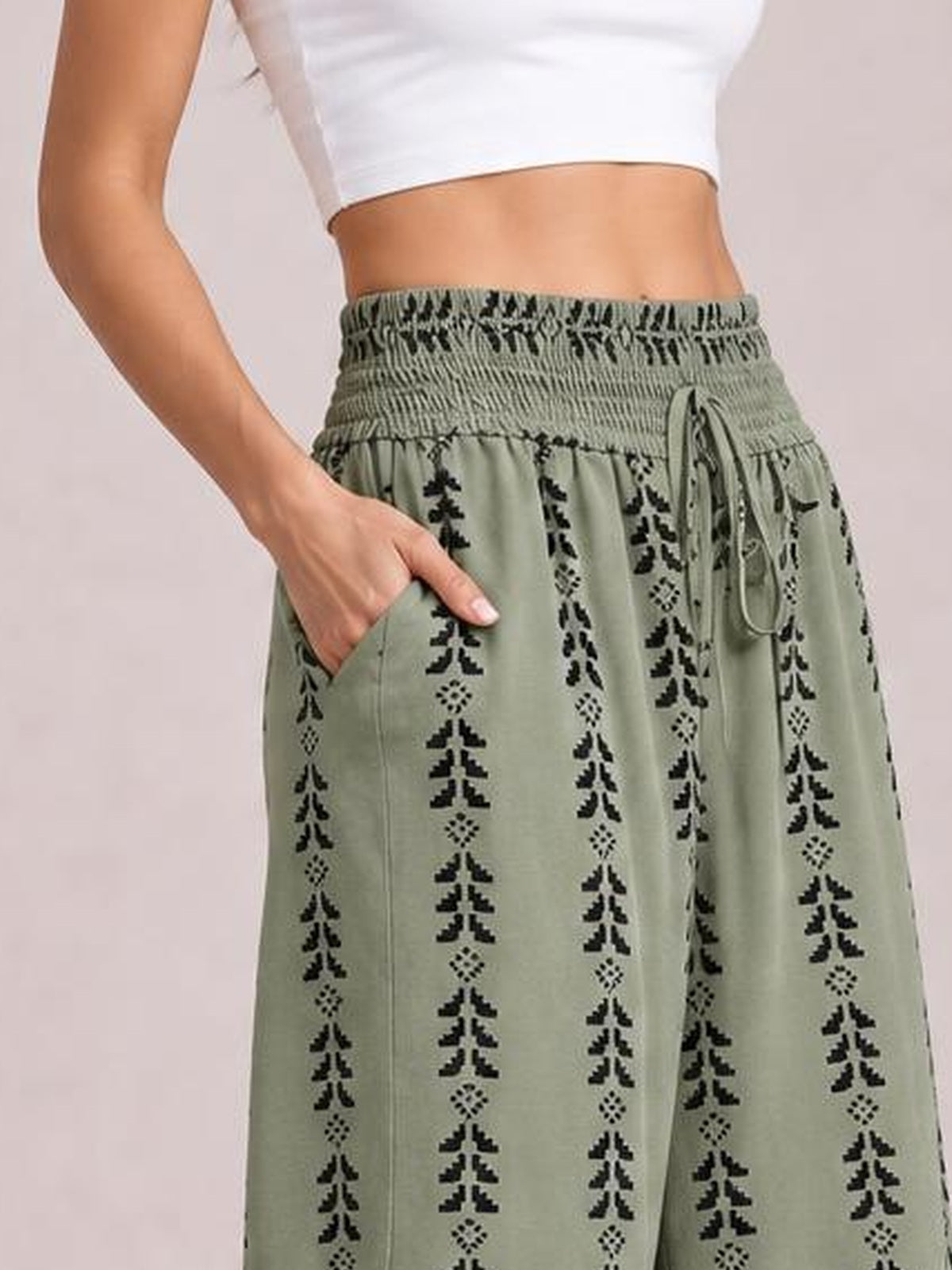 A model wears sage green tied printed high-waist pants with a smocked waistband, drawstring, vertical black motifs, wide-leg silhouette, and a side pocket.