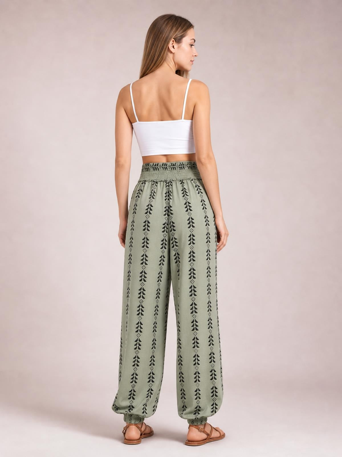 A model poses from behind in sage green printed high-waist pants with a smocked waistband, relaxed wide legs, and elasticized cuffs.