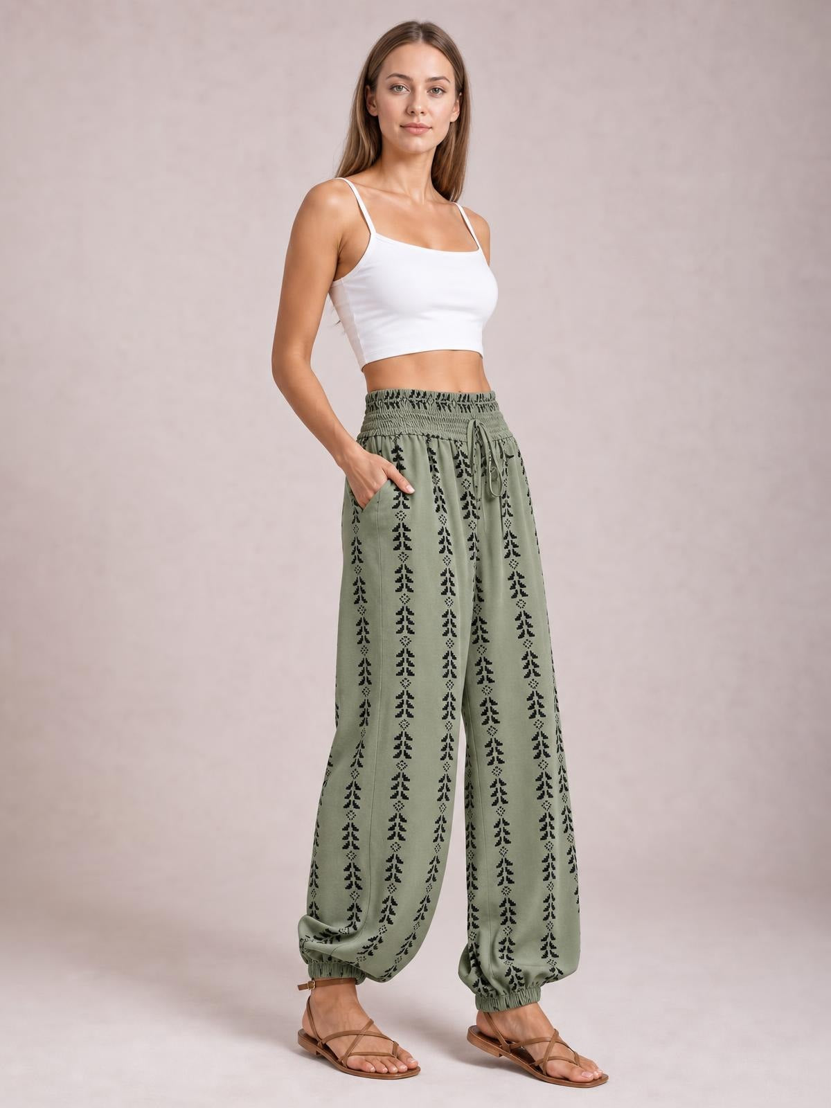 Model poses in high-waist sage green printed pants with a wide-leg silhouette, elastic cuffs, gathered waistband, side pockets, and a tied drawstring.