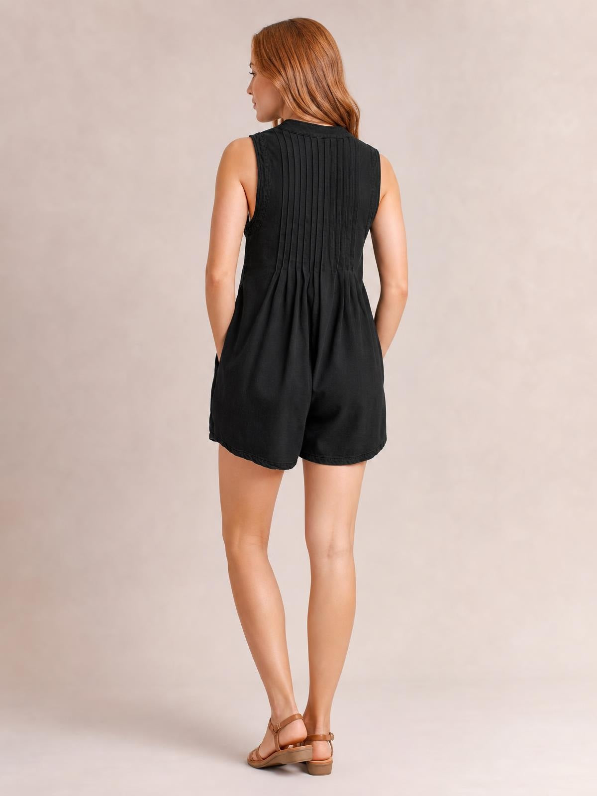 Model shown from behind in a black sleeveless romper with a high neckline, vertical pleats, relaxed shorts silhouette, and side pockets.