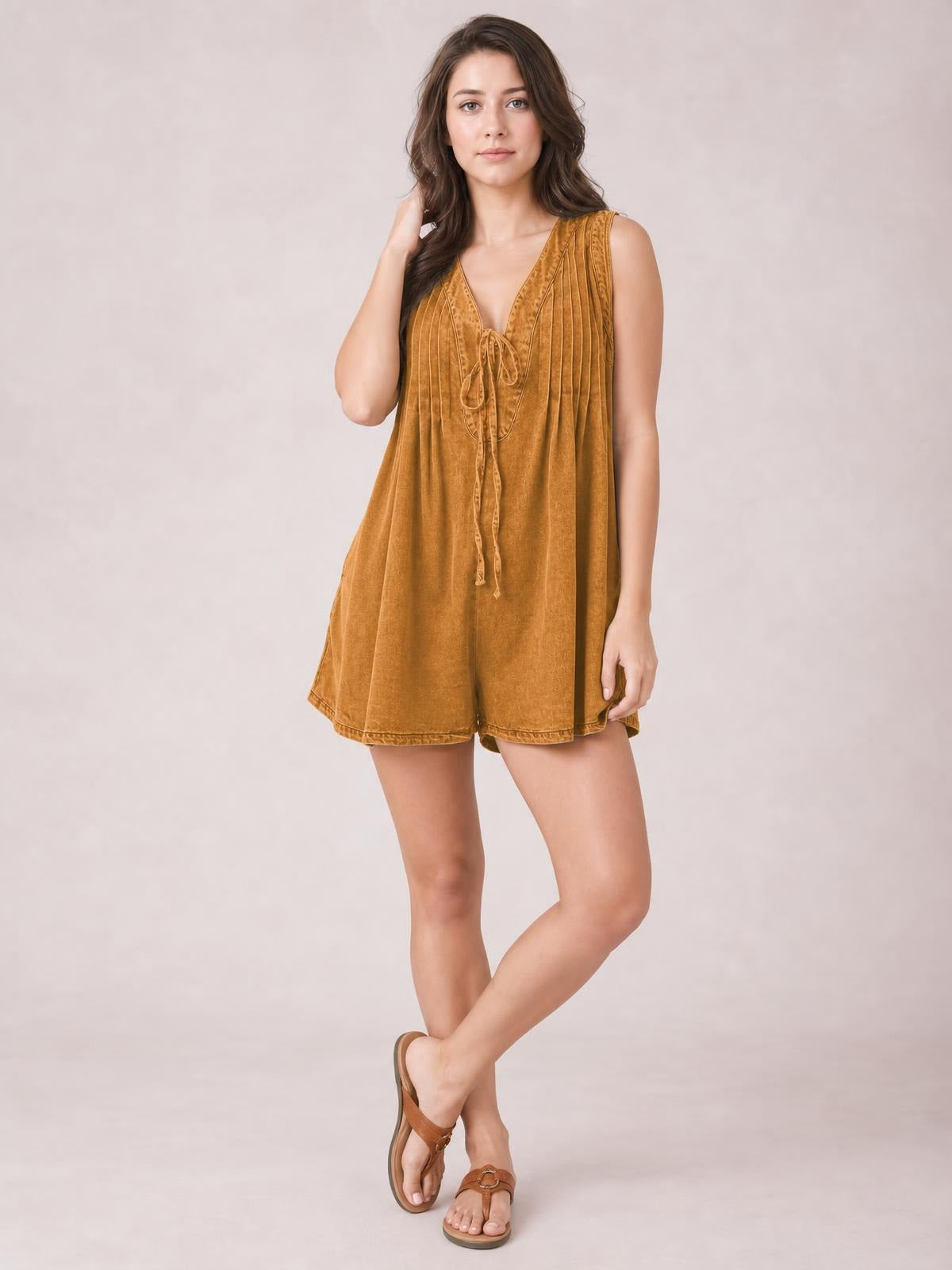 A model poses with crossed legs in a mustard-brown sleeveless romper featuring a V-neck, tied front, vertical pleats, and a relaxed shorts silhouette.