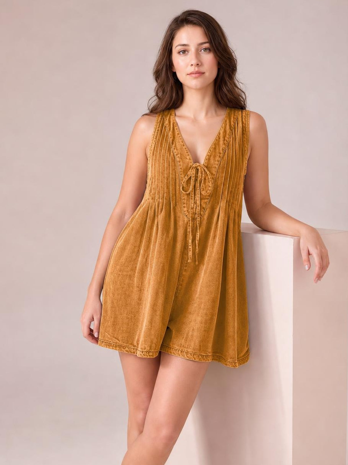 A model poses in a mustard-gold tied romper with a sleeveless deep V-neck, softly flared short silhouette, vertical panel construction, and practical pockets.