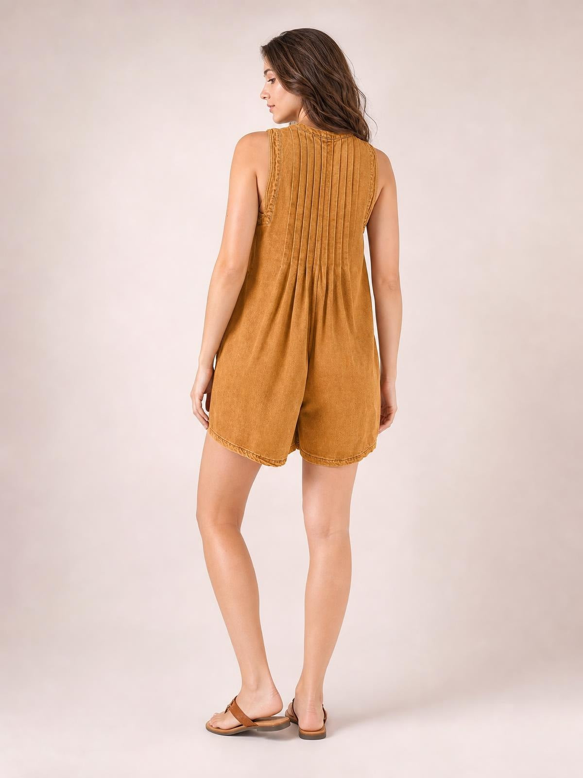 A model shows the back of a caramel brown sleeveless romper with vertical pleats, a relaxed shorts silhouette, and neatly finished hems.