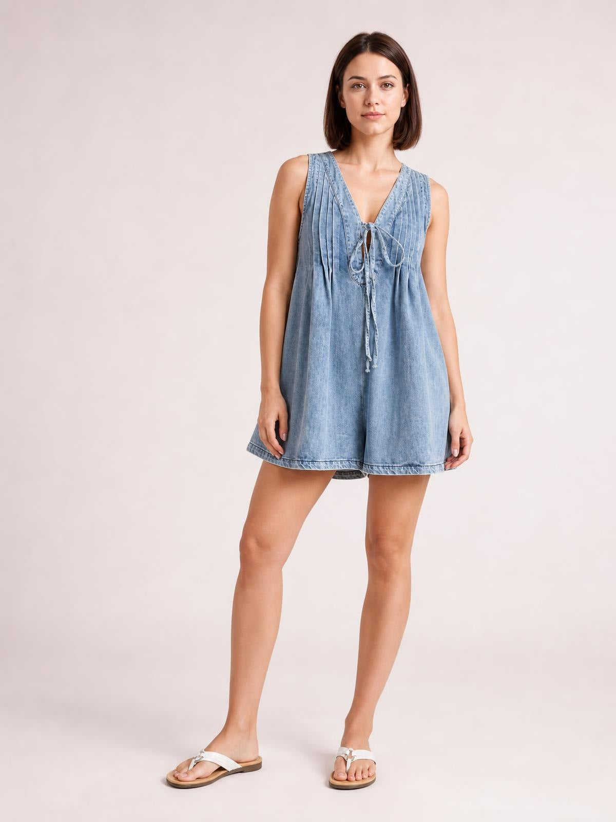 A model stands in a light blue sleeveless romper with a V-neck, front tie detail, relaxed wide-leg shorts, and practical pockets.