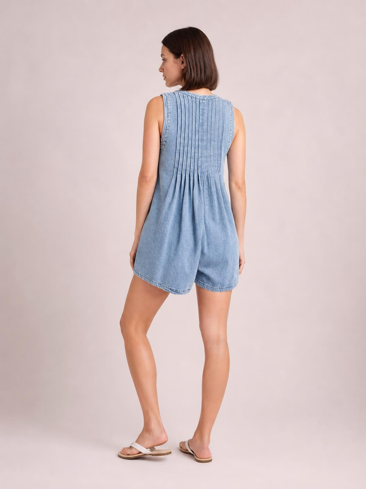 A model poses turned away in a light blue sleeveless romper with a round neckline, pleated back construction, relaxed shorts silhouette, and stitched hems.