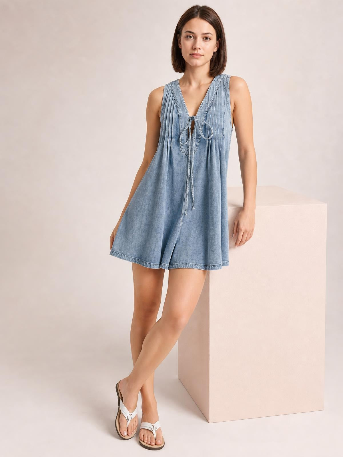 Model in a light blue denim romper with a tied V-neck, sleeveless design, pleated construction, relaxed silhouette, and shorts-length hem.