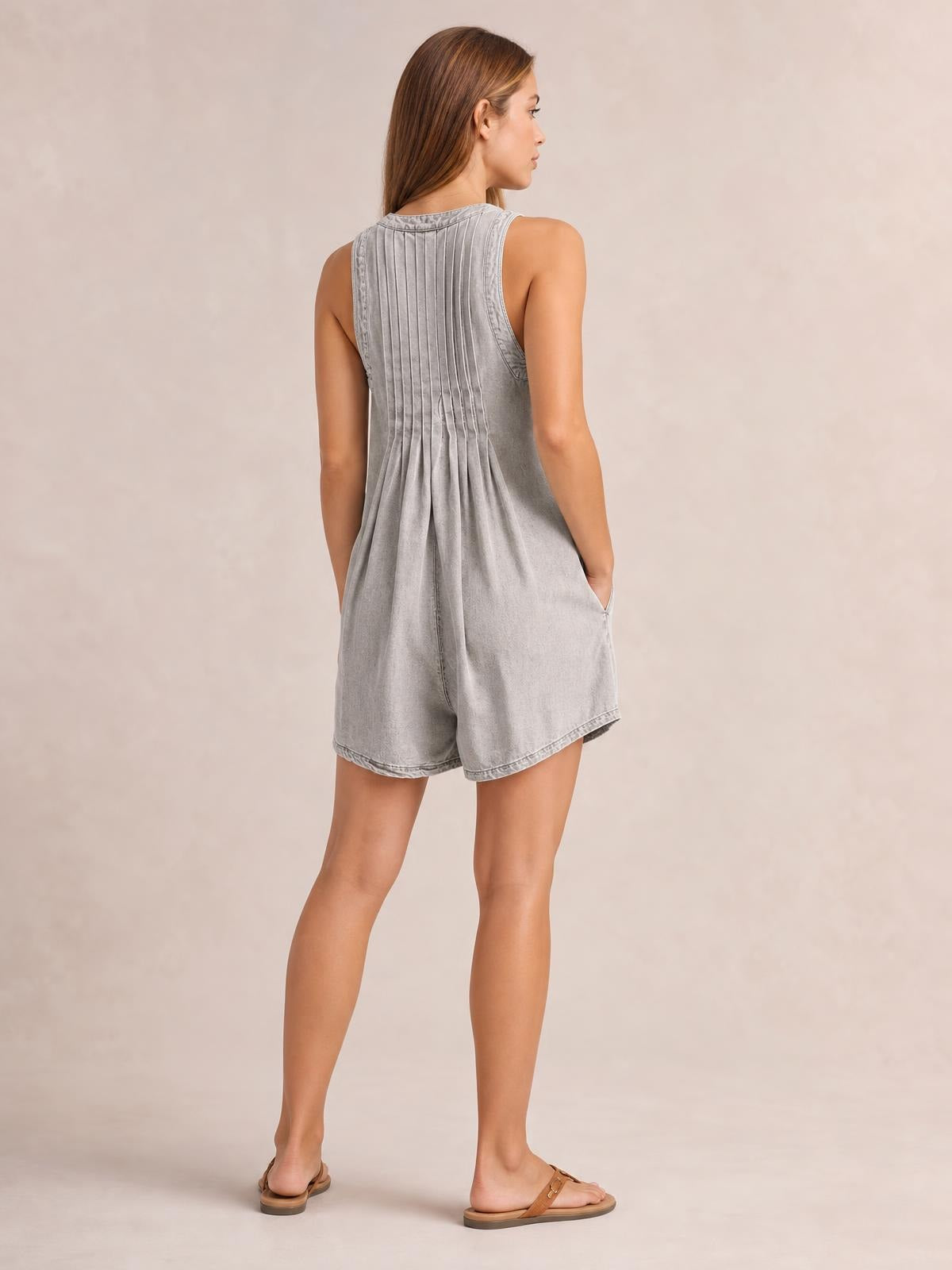 Model wearing a light gray sleeveless romper with a pleated back, relaxed shorts silhouette, and side pockets, posing with one hand tucked inside.
