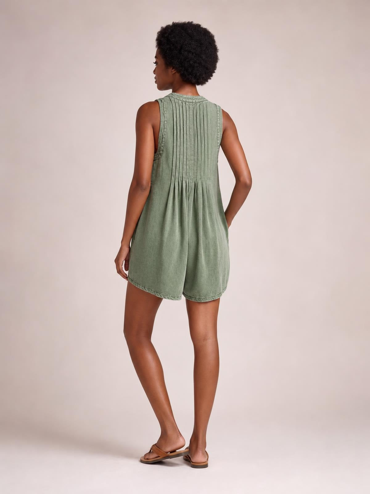 A model turns to show a sage-green sleeveless romper with a relaxed shorts silhouette, pleated back construction, and side pockets.