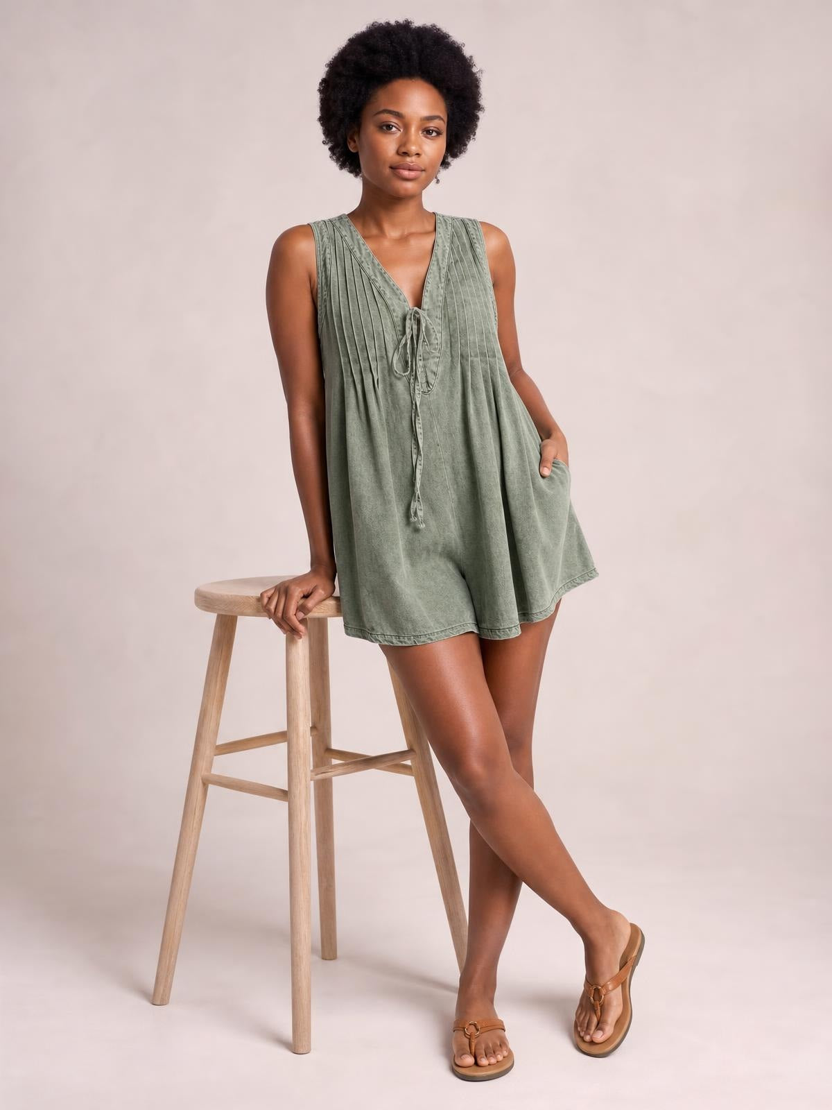 A model wears an olive green sleeveless romper with a V-neck, tied front, pleated bodice, relaxed short silhouette, and side pockets while leaning on a stool.