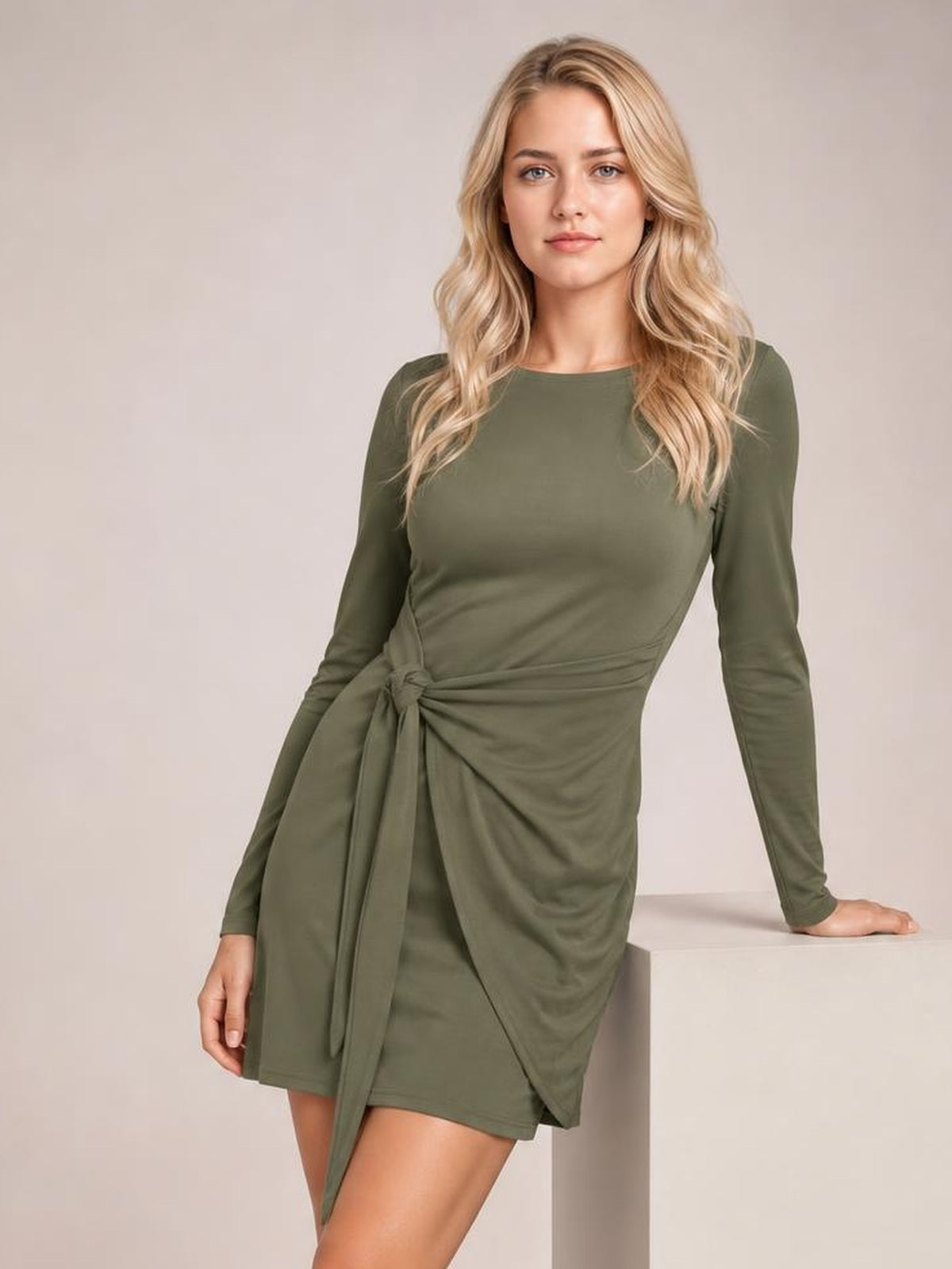 A model poses in an olive green tied round-neck mini dress with long sleeves, a fitted silhouette, gathered waist, and long draped sash.