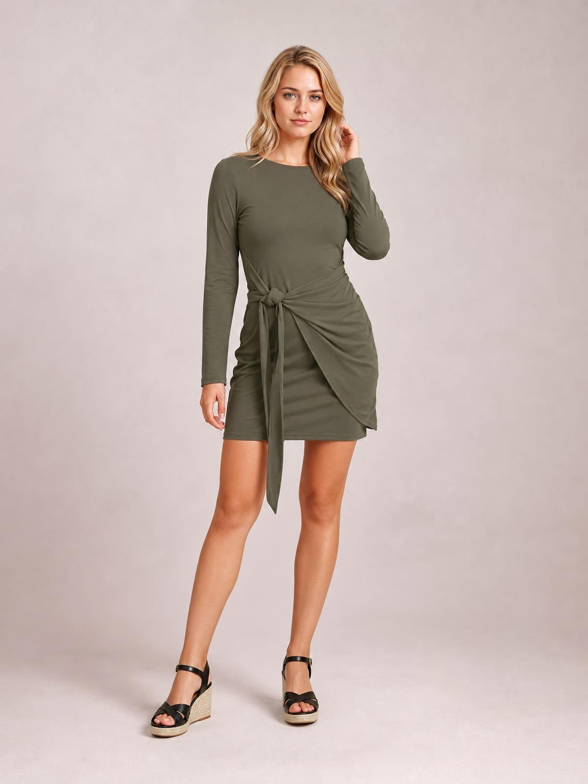Model wearing an olive green long sleeve mini dress with a round neck, fitted silhouette, and tied ruched waist detail with a draped overlay.
