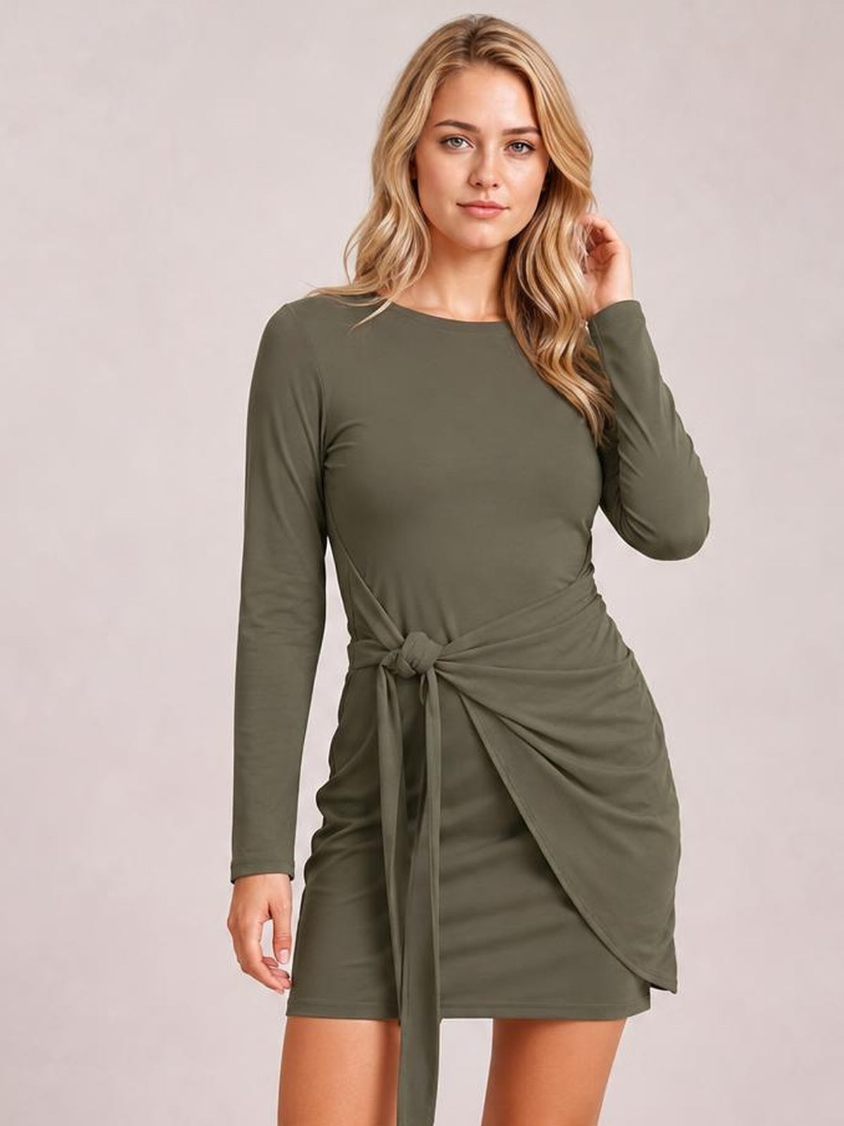 Model wears an olive green round-neck long-sleeve mini dress with a fitted silhouette and tied waist detail, posing with one hand near her hair.