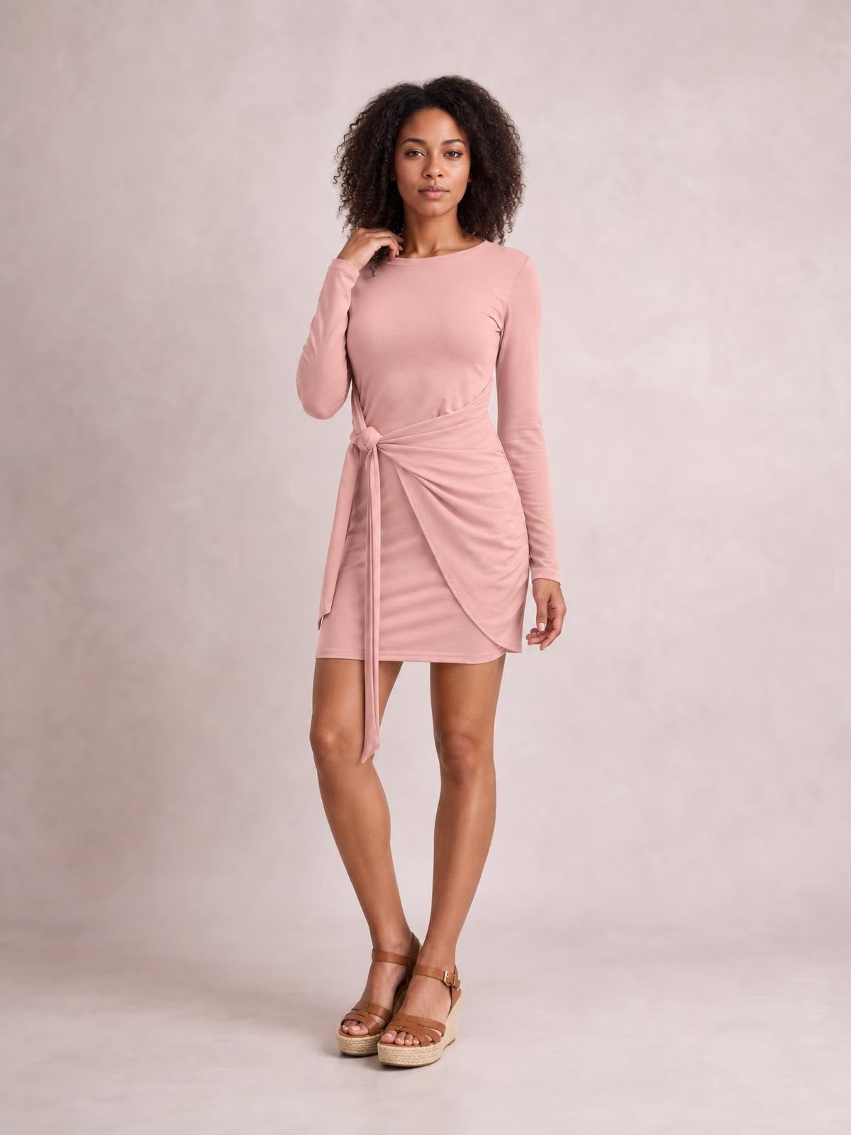 Model poses in a blush pink tied round-neck long-sleeve mini dress with a fitted silhouette and draped wrap-style waist detail.