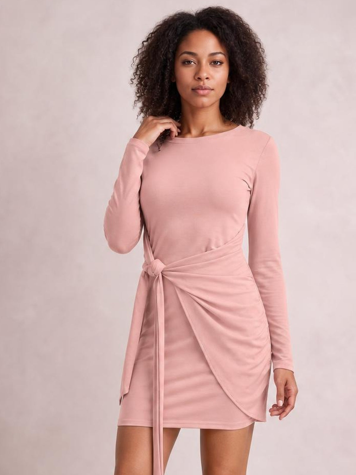 Model wearing a blush pink round neck long sleeve mini dress with a tied wrap waist, draped skirt panel, and fitted silhouette.