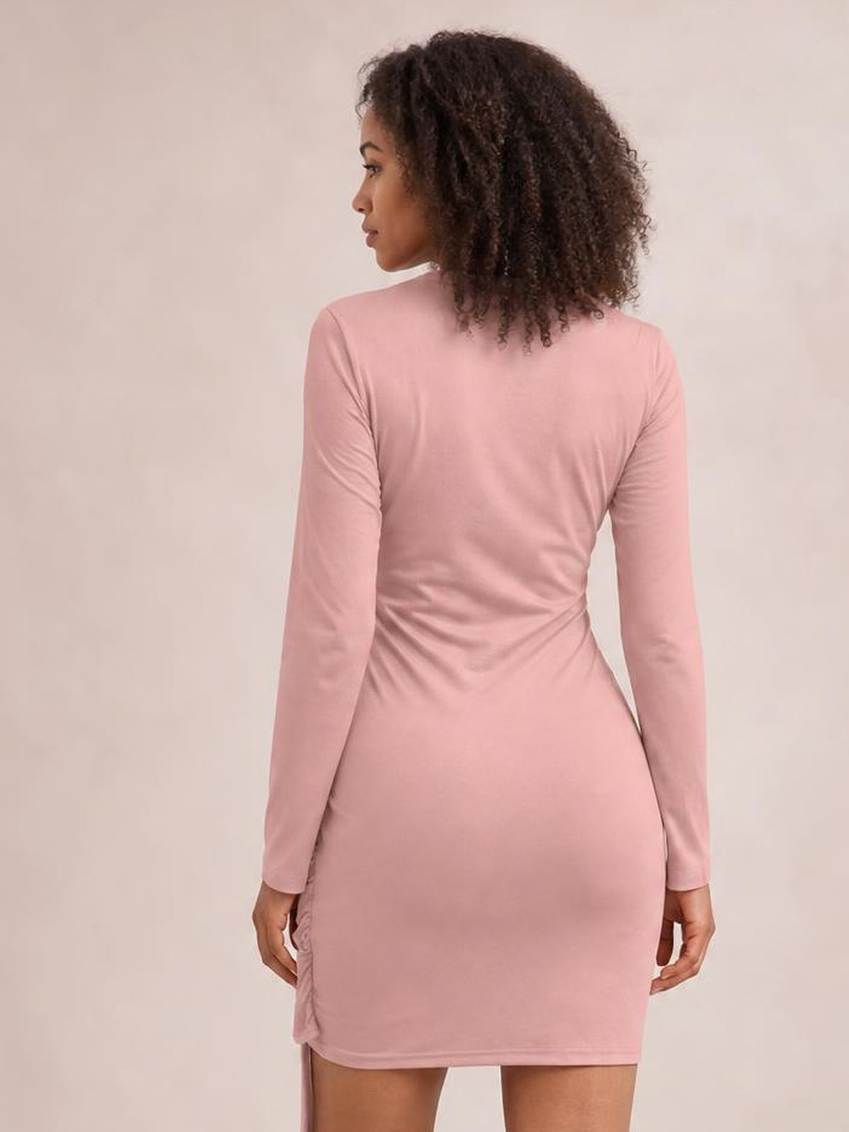 A model looks over her shoulder wearing a blush pink tied round-neck mini dress with long sleeves, a fitted silhouette, and gathered side detailing.
