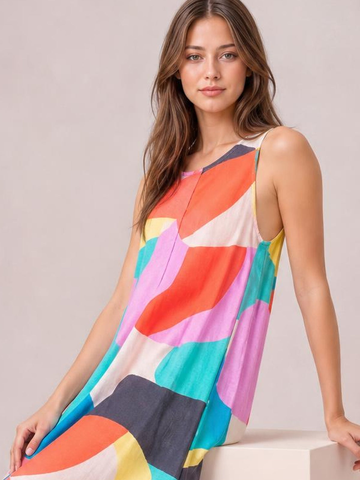 Model wears a colorful geometric-print jumpsuit with shoulder straps and a relaxed sleeveless silhouette, leaning casually with one hand resting beside her.