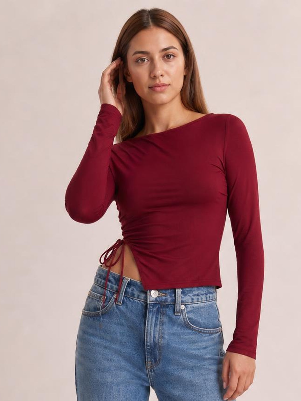 A model poses in a burgundy long-sleeve T-shirt with a round neck, fitted cropped silhouette, and tied side slit creating ruched detailing.