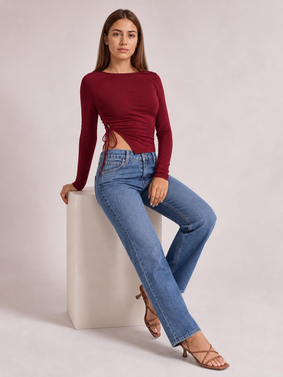 Model seated in a burgundy tied slit round neck long sleeve T-shirt with a fitted silhouette, ruched waist, and adjustable side ties.