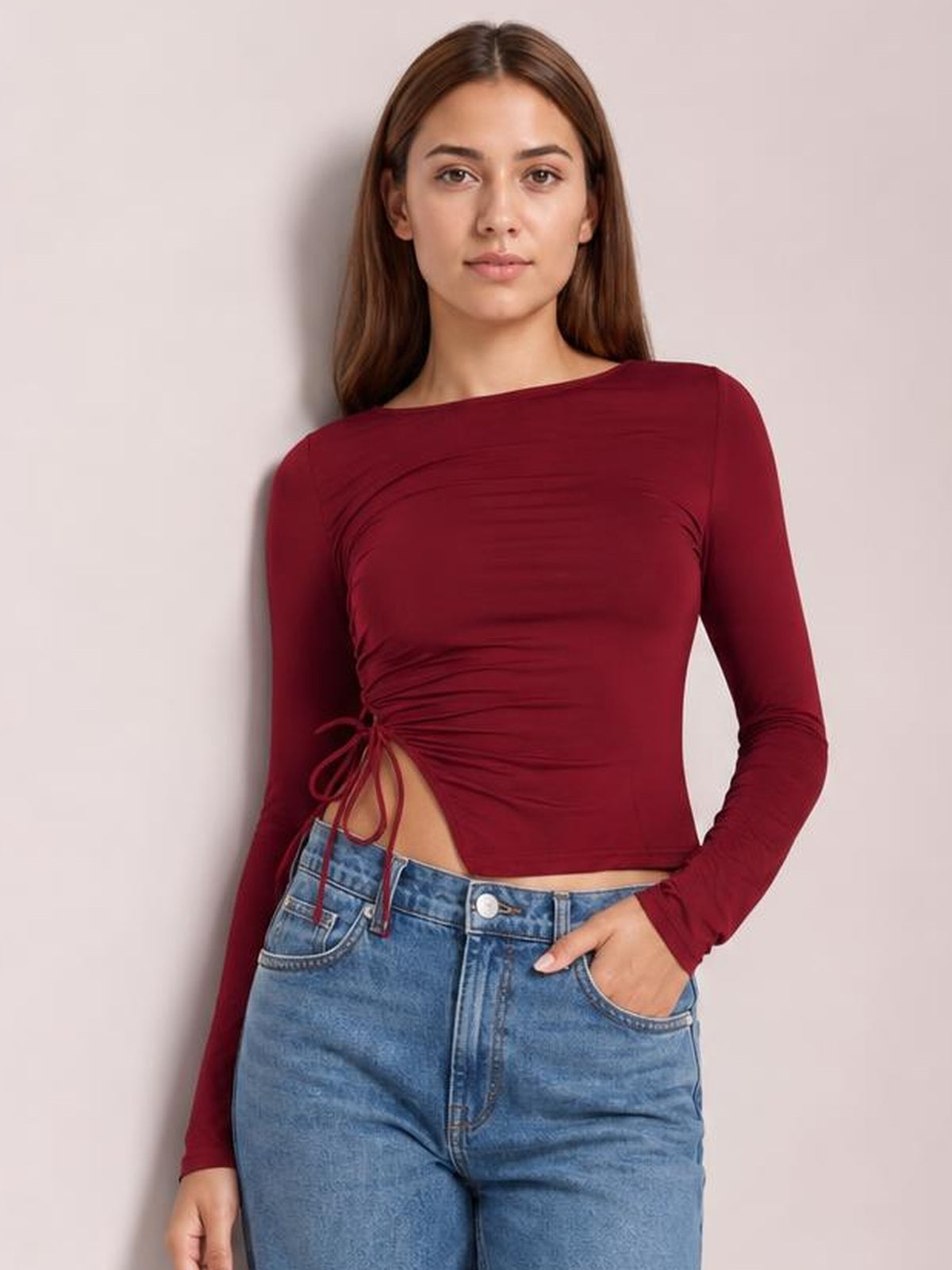 Model wearing a burgundy tied slit round-neck long-sleeve T-shirt with a fitted cropped silhouette and ruched side detail, posing with one hand in her jeans pocket.