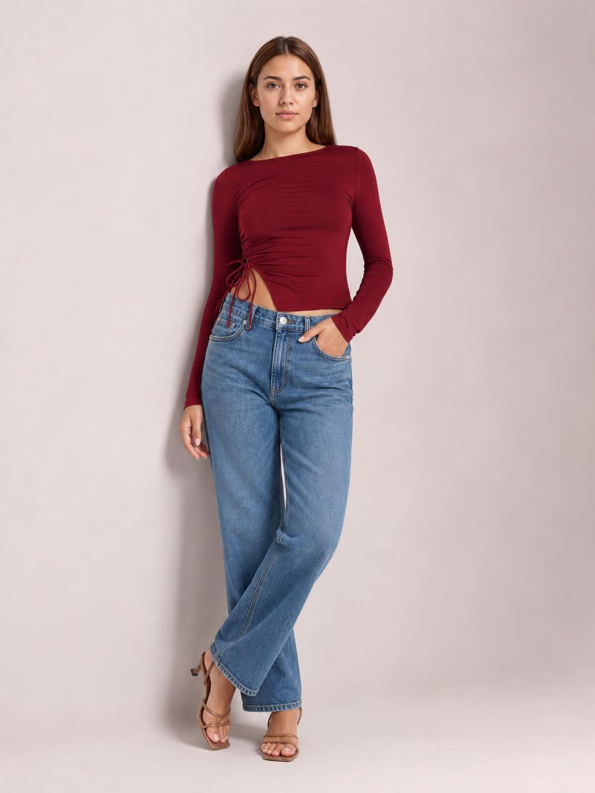 A model poses in a burgundy tied slit round-neck long-sleeve T-shirt with a ruched, fitted cropped silhouette and side cutout.
