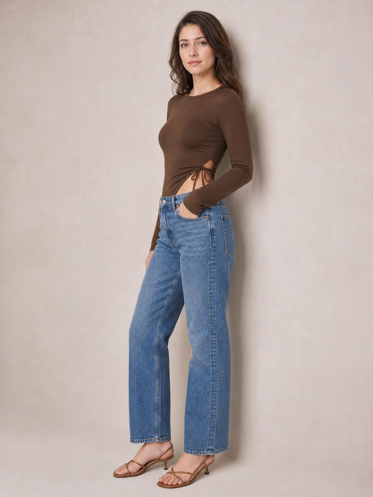 Model wears a fitted brown round-neck long-sleeve T-shirt with a tied side slit detail, posing with one hand in her jeans pocket.
