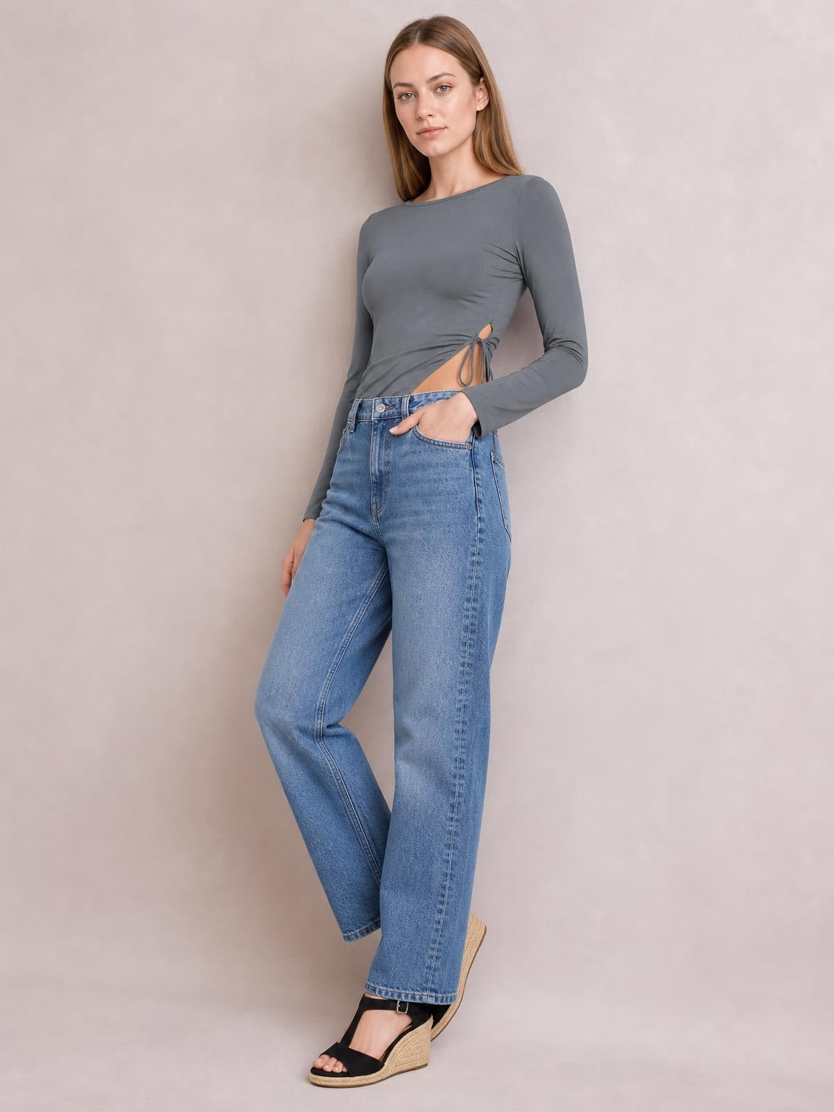Model poses in a gray long-sleeve round-neck T-shirt with a tied side slit, fitted silhouette, and ruched waist detail.