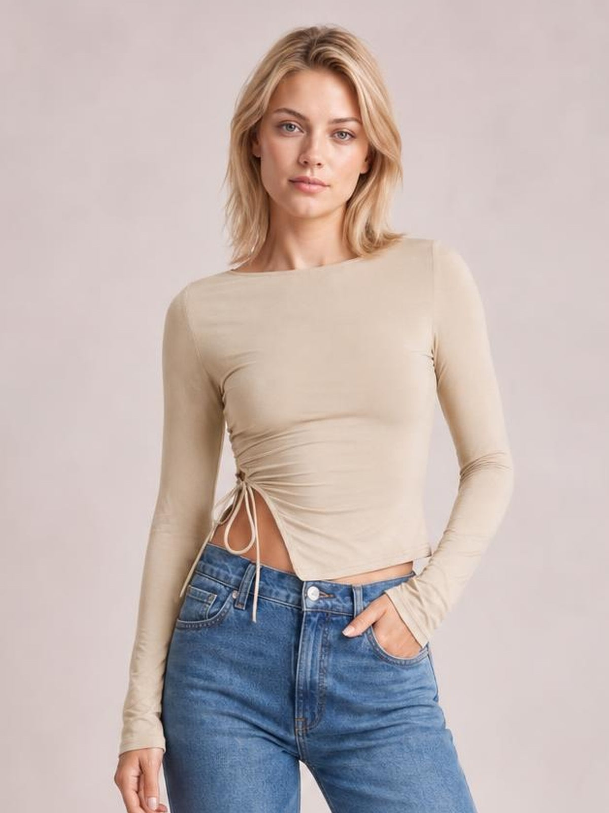 Model wears a fitted beige long-sleeve T-shirt with a round neck, side slit and tied detail creating a ruched waist, posing with one hand in her jeans pocket.