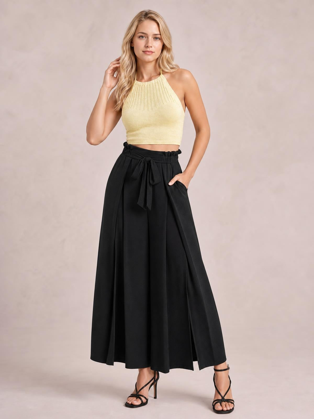 A model poses in black tied slit wide-leg pants with a high waist, flowing floor-length silhouette, visible pockets, and one hand tucked inside.